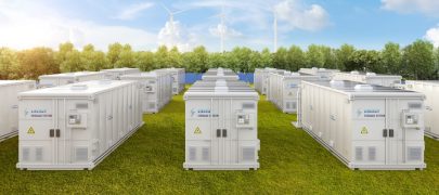 Large commercial solar energy batteries surrounded by green fields and blue skies with wind turbines in the background